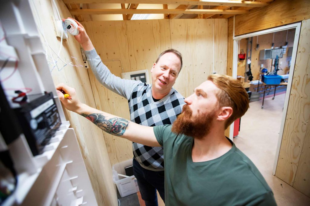 Två män - en med rött skägg och tatuering på underarmen, en med stickad tröja, står mot vägg och jobbar med elektronisk utrustning.